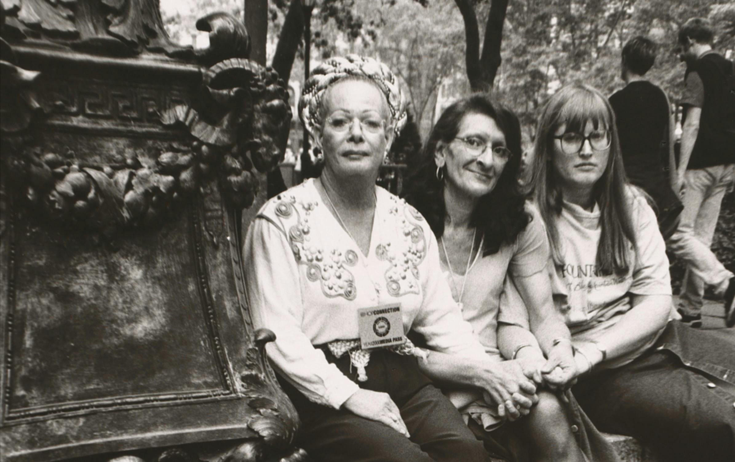 Sylvia Rivera, Christina Hayworth and Julia Murray sit on a ledge in a park in New York City. A sign near them reads: Respect Trans People/Men!