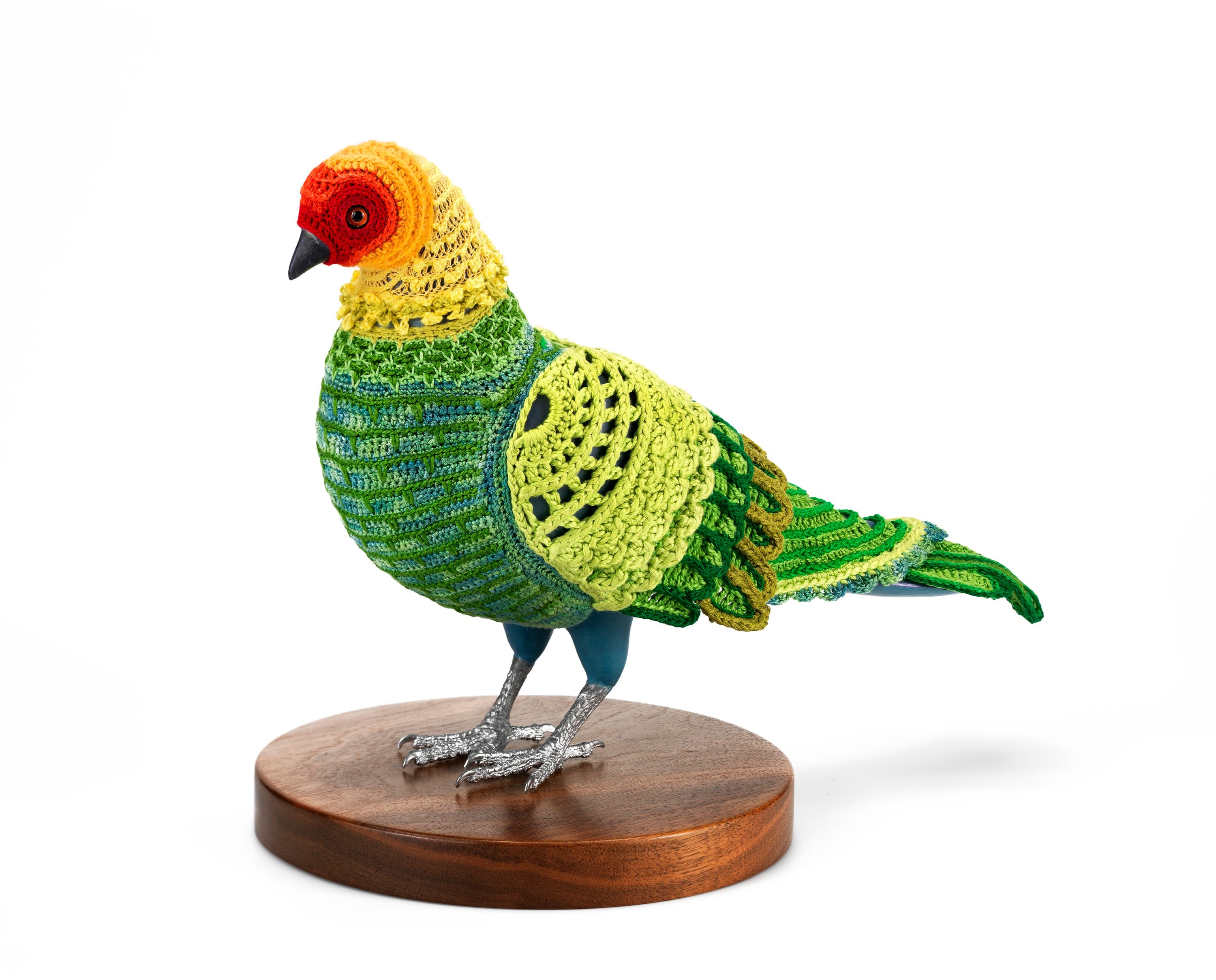 A crocheted suit with green, yellow, and red patterns on a pigeon mannequin, standing on a round wooden base