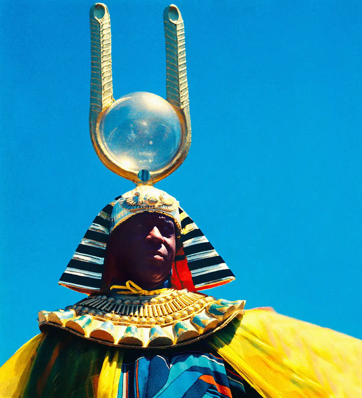 A person wearing a colorful flowing costume with a striped headdress with a clear orb supported by golden horn-like prongs